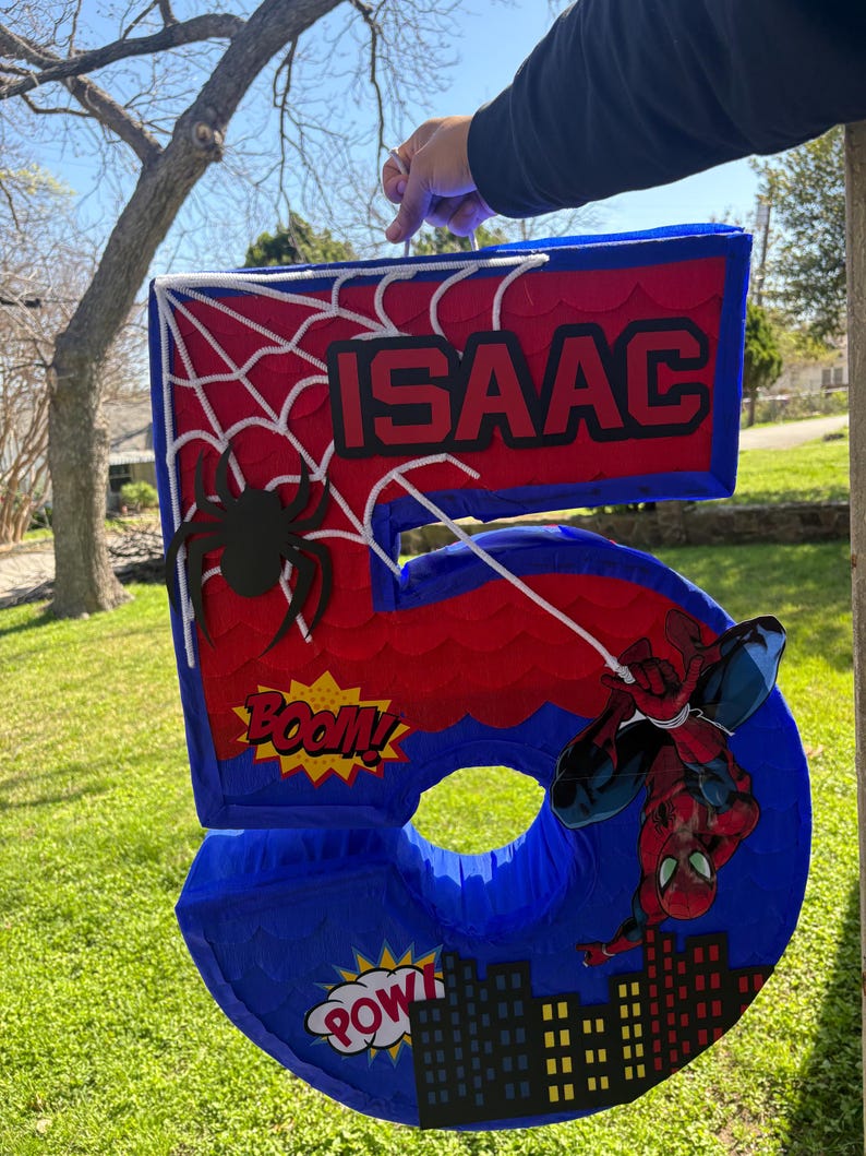 K&ouml;nnte beinhalten: Eine blaue, rote und wei&szlig;e Pi&ntilde;ata mit der Nummer 5 im Spider-Man-Design. Die Pi&ntilde;ata hat ein Spinnennetz-Design, eine schwarze Spinne und den Namen "ISAAC" in Rot. Comic-Soundeffekte und eine Skyline sind ebenfalls enthalten.