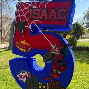 K&ouml;nnte beinhalten: Eine gro&szlig;e Pi&ntilde;ata in Form der Zahl 5 in Blau, Rot und Wei&szlig;, mit einem Spider-Man-Thema. Die Pi&ntilde;ata enth&auml;lt den Namen "ISAAC" und Comic-Textblasen mit "BOOM!" und "POW!". Eine Spider-Man-Figur ist angebracht.