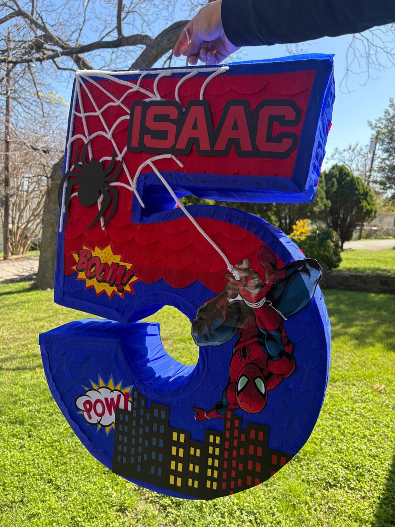 K&ouml;nnte beinhalten: Eine gro&szlig;e Pi&ntilde;ata in Form der Zahl 5 in Blau, Rot und Wei&szlig;, mit einem Spider-Man-Thema. Die Pi&ntilde;ata enth&auml;lt den Namen "ISAAC" und Comic-Textblasen mit "BOOM!" und "POW!". Eine Spider-Man-Figur ist angebracht.