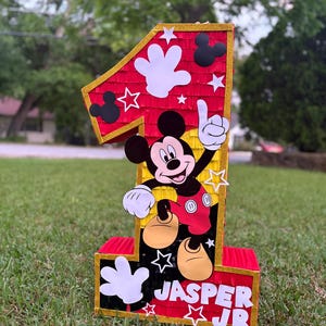 May include: A large, number one pi&ntilde;ata featuring Mickey Mouse. The pi&ntilde;ata is red with gold trim and white star and hand accents. The name "JASPER JR" is displayed on the base. The pi&ntilde;ata is on a grassy lawn.