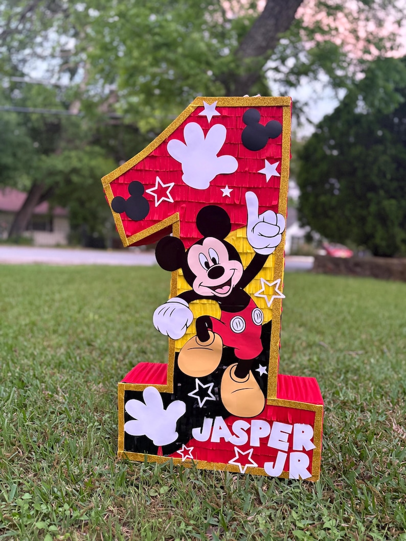 May include: A large, number one pi&ntilde;ata featuring Mickey Mouse. The pi&ntilde;ata is red with gold trim and white star and hand accents. The name "JASPER JR" is displayed on the base. The pi&ntilde;ata is on a grassy lawn.