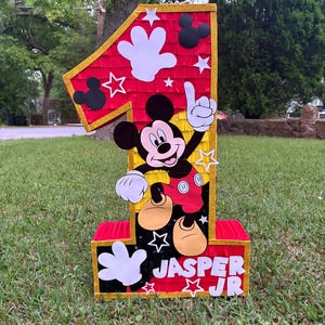 May include: A large, number one pi&ntilde;ata featuring Mickey Mouse. The pi&ntilde;ata is red, yellow, and black with white accents, including Mickey's gloves and stars. The name "Jasper Jr" is printed on the base.