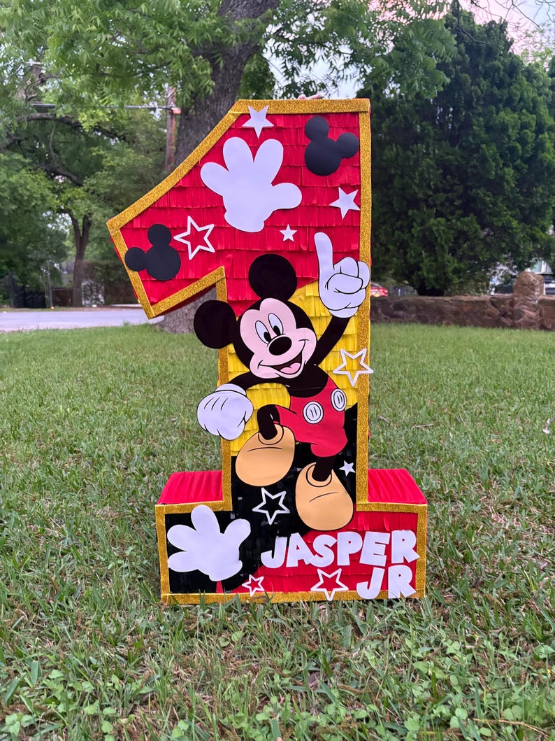 May include: A large, number one pi&ntilde;ata featuring Mickey Mouse. The pi&ntilde;ata is red, yellow, and black with white accents, including Mickey's gloves and stars. The name "Jasper Jr" is printed on the base.