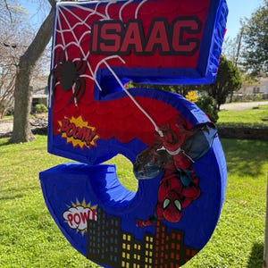 K&ouml;nnte beinhalten: Eine gro&szlig;e, blaue, rote und schwarze Pi&ntilde;ata mit der Zahl 5 und einem Spider-Man-Motiv. Die Pi&ntilde;ata tr&auml;gt den Namen "ISAAC" und Comic-Soundeffekte. Eine schwarze Spinne und eine Stadtlandschaft sind ebenfalls zu sehen.