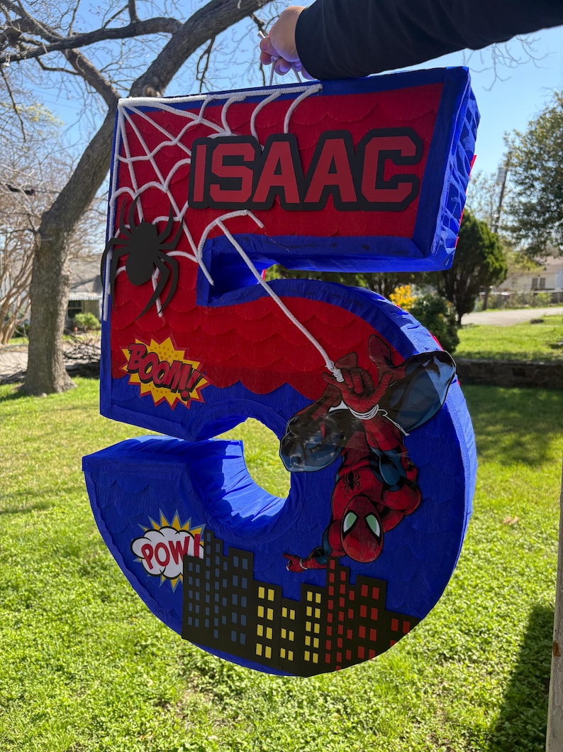K&ouml;nnte beinhalten: Eine gro&szlig;e, blaue, rote und schwarze Pi&ntilde;ata mit der Zahl 5 und einem Spider-Man-Motiv. Die Pi&ntilde;ata tr&auml;gt den Namen "ISAAC" und Comic-Soundeffekte. Eine schwarze Spinne und eine Stadtlandschaft sind ebenfalls zu sehen.