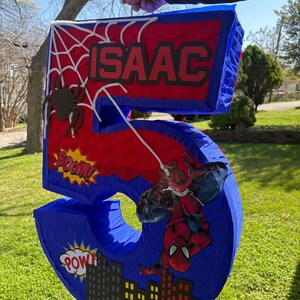 K&ouml;nnte beinhalten: Eine blaue und rote Zahl 5 Pi&ntilde;ata mit Spider-Man-Motiv. Die Pi&ntilde;ata zeigt den Namen "ISAAC" in wei&szlig;en Buchstaben, ein Spinnennetz und eine Spider-Man-Grafik. Comic-Soundeffekte "BOOM!" und "POW!" sind ebenfalls enthalten.