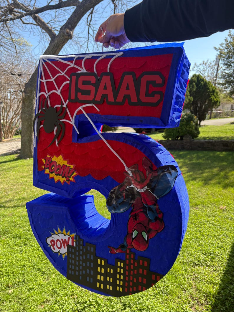 K&ouml;nnte beinhalten: Eine blaue und rote Zahl 5 Pi&ntilde;ata mit Spider-Man-Motiv. Die Pi&ntilde;ata zeigt den Namen "ISAAC" in wei&szlig;en Buchstaben, ein Spinnennetz und eine Spider-Man-Grafik. Comic-Soundeffekte "BOOM!" und "POW!" sind ebenfalls enthalten.