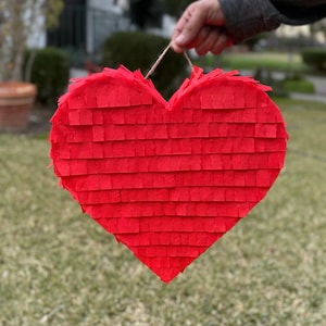 May include: A large, heart-shaped piñata made of red crepe paper. The piñata is held by a string and is against a background of green grass and a blurred residential area.