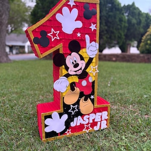 May include: A large, red number one pi&ntilde;ata with gold trim, Mickey Mouse graphics, and white star and hand cutouts. The pi&ntilde;ata has the name "JASPER JR" printed on the front. The pi&ntilde;ata is sitting on green grass.