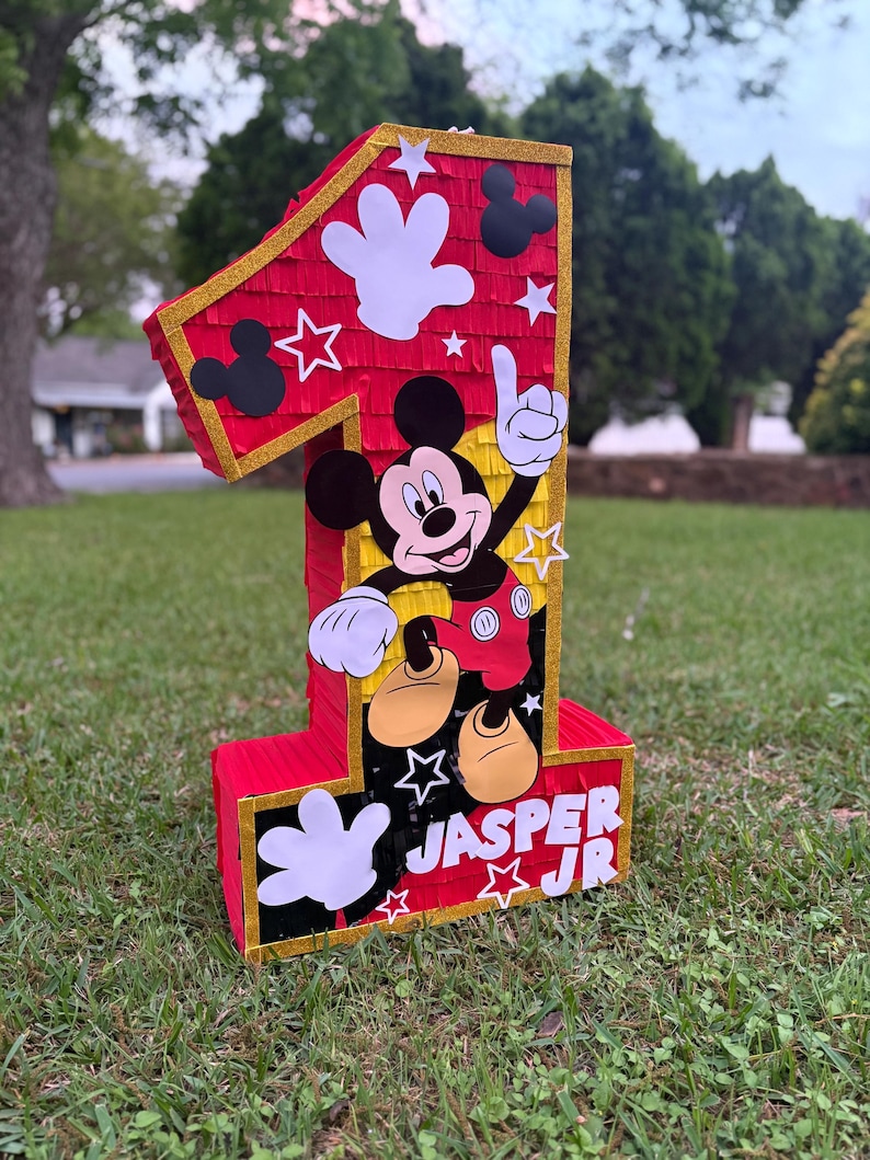 May include: A large, red number one pi&ntilde;ata with gold trim, Mickey Mouse graphics, and white star and hand cutouts. The pi&ntilde;ata has the name "JASPER JR" printed on the front. The pi&ntilde;ata is sitting on green grass.