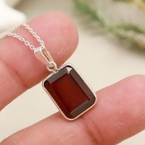 May include: A silver necklace featuring a rectangular, dark red gemstone pendant. The pendant is set in a silver frame and hangs from a delicate silver chain. The gemstone has a polished, reflective surface, and the overall design is simple and elegant.