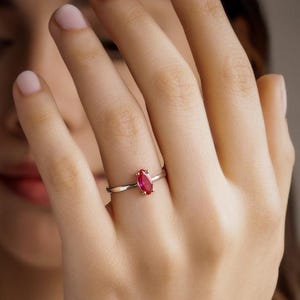 May include: A silver ring with a marquise-cut, bright red gemstone. The ring is simple and elegant, with a thin band. The gemstone is the focal point, catching the light and adding a pop of color. The ring is shown on a hand.