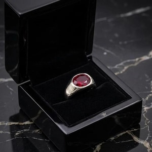 Puede incluir: Un anillo de plata con una gran gema roja oscura de corte ovalado, presentado en una caja de joyería negra con interior de terciopelo. La caja está abierta, revelando la banda de metal pulido del anillo y el color vibrante de la piedra.