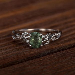 May include: A silver ring with a green oval gemstone. The ring features a leaf design with small accent stones. The ring is set against a dark wood surface, highlighting the ring's details and color.