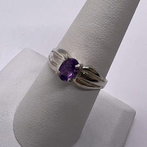May include: A silver ring featuring an oval-cut, deep purple gemstone. The ring has a simple, elegant design with a split shank and a polished finish. The gemstone is set in a classic prong setting, showcasing its color and clarity.