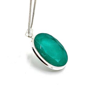 Peut inclure: Un collier pendentif en argent avec une grosse pierre précieuse verte émeraude de forme ovale. La pierre est sertie dans une lunette en argent et suspendue à une fine chaîne en argent. Le pendentif est présenté sur fond blanc.
