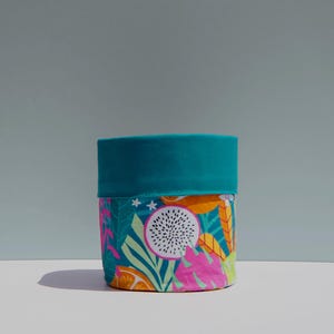 May include: A cylindrical container featuring a teal rim and a body decorated with a colourful tropical fruit and leaf pattern. The design incorporates pink, orange, and white elements against a teal backdrop. The container rests on a white surface.