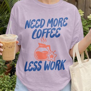 Puede incluir: Camiseta lila con la frase azul "NEED MORE COFFEE LESS WORK" y un diseño de cafetera. La persona sostiene una bebida de café y una pequeña bolsa de mano. También se ven unos pantalones cortos a rayas azules y blancas.