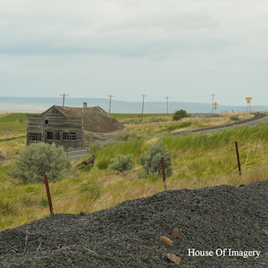 May include: A weathered, wooden building with a faded roof stands in a grassy field under a cloudy sky. A road curves through the landscape, with power lines and a fence in the foreground. The scene evokes a sense of rural isolation.