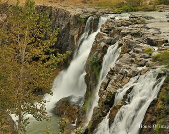 May include: A scenic view of a waterfall cascading over rocky cliffs. The water flows rapidly, creating a white, foamy effect against the dark, textured rocks. Green foliage and trees frame the waterfall, adding natural color.