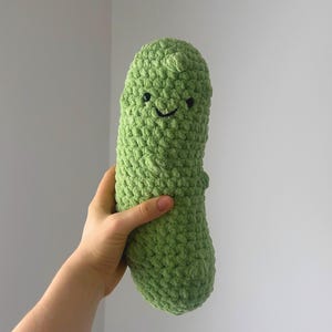 May include: A handmade, green crochet pickle plushie with a smiling face. The plush toy is held by a hand against a plain background. The pickle has a textured surface and a rounded shape.