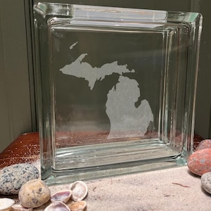 May include: A clear glass block etched with the outline of the state of Michigan. The block is square and sits on a surface with sand, seashells, and stones. The glass block is a decorative item.