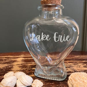 May include: A clear, heart-shaped glass bottle with a cork stopper and the words "Lake Erie" in white script. Several seashells are scattered near the bottle. The bottle is on a wooden surface.