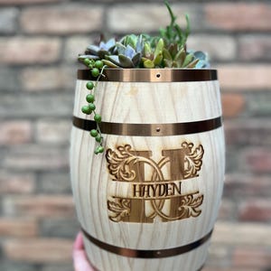 May include: A wooden barrel planter with a copper-colored metal trim and the name "HAYDEN" engraved on the front. The planter is filled with a variety of succulents and trailing plants. The barrel is held by a person.