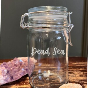 May include: A clear glass jar with a hinged lid and metal clasp. The words "Dead Sea" are written in a cursive font on the front of the jar. The jar is sitting on a wooden surface.