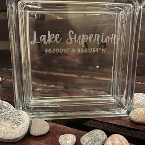 May include: A clear glass block etched with "Lake Superior" and coordinates 46.70015° N 85.41754° W. The block is square and sits on a wooden surface with smooth stones in the foreground. A decorative item.