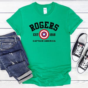 May include: A green t-shirt with the text "ROGERS EST 1918 CAPTAIN AMERICA" in black. The shirt features a Captain America shield graphic. Dark blue jeans and black and white sneakers are also in the image.