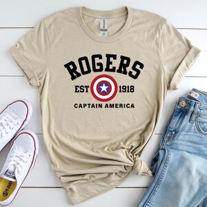 May include: A tan t-shirt with the text "ROGERS EST 1918 CAPTAIN AMERICA" in black, red, and white. The shirt is paired with blue jeans and white sneakers with red trim.