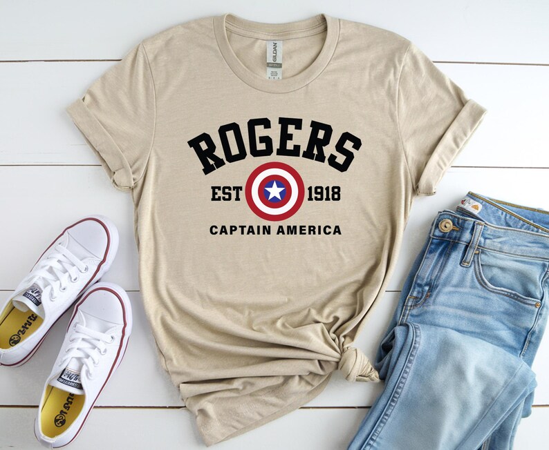 May include: A tan t-shirt with the text "ROGERS EST 1918 CAPTAIN AMERICA" in black, red, and white. The shirt is paired with blue jeans and white sneakers with red trim.