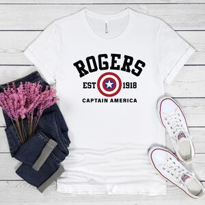 May include: White t-shirt with the text "ROGERS" above a Captain America shield graphic. Below the graphic are the words "EST 1918 CAPTAIN AMERICA" in black. The shirt is paired with blue jeans and white sneakers.
