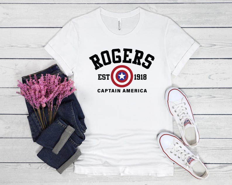 May include: White t-shirt with the text "ROGERS" above a Captain America shield graphic. Below the graphic are the words "EST 1918 CAPTAIN AMERICA" in black. The shirt is paired with blue jeans and white sneakers.