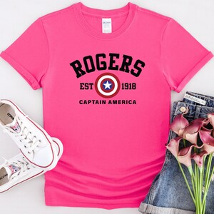 May include: A bright pink t-shirt with the text "ROGERS EST 1918 CAPTAIN AMERICA" in black. The design includes a Captain America shield graphic. White sneakers and denim shorts are also in the image.
