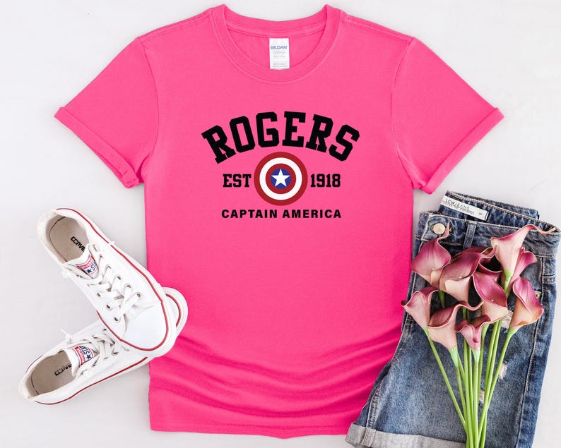 May include: A bright pink t-shirt with the text "ROGERS EST 1918 CAPTAIN AMERICA" in black. The design includes a Captain America shield graphic. White sneakers and denim shorts are also in the image.
