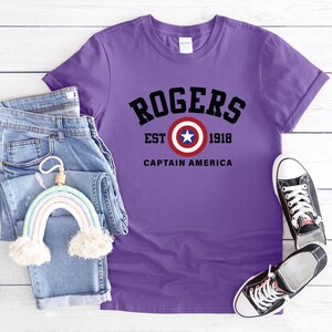 May include: A purple t-shirt with the text "ROGERS EST 1918 CAPTAIN AMERICA" in black. The shirt is paired with blue jeans, a rainbow-shaped decoration, and black and white sneakers.