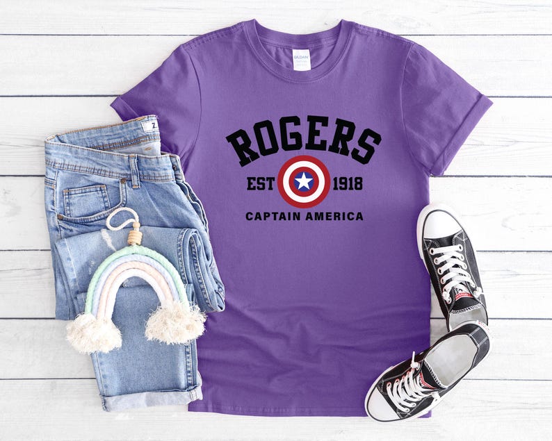 May include: A purple t-shirt with the text "ROGERS EST 1918 CAPTAIN AMERICA" in black. The shirt is paired with blue jeans, a rainbow-shaped decoration, and black and white sneakers.