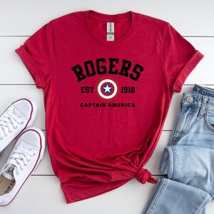 May include: A red t-shirt with the text "ROGERS EST 1918 CAPTAIN AMERICA" in black. The shirt is paired with light blue jeans and white sneakers. The design includes a shield with a star.
