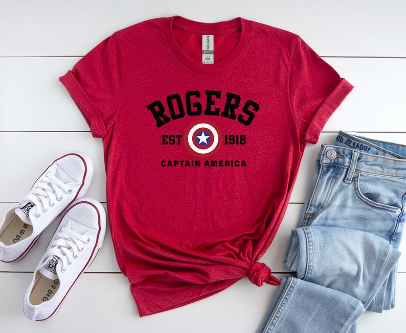 May include: A red t-shirt with the text "ROGERS EST 1918 CAPTAIN AMERICA" in black. The shirt is paired with light blue jeans and white sneakers. The design includes a shield with a star.