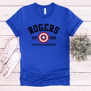 May include: Royal blue t-shirt with the text "ROGERS EST 1918 CAPTAIN AMERICA" in black and white. The design includes a circular emblem with a white star on a red and white background. White canvas sneakers are on the left.