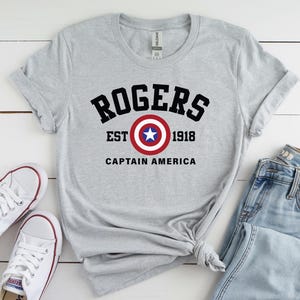 May include: A gray t-shirt with the text "ROGERS EST 1918 CAPTAIN AMERICA" in black and a Captain America shield graphic. The shirt is paired with white sneakers and blue jeans.