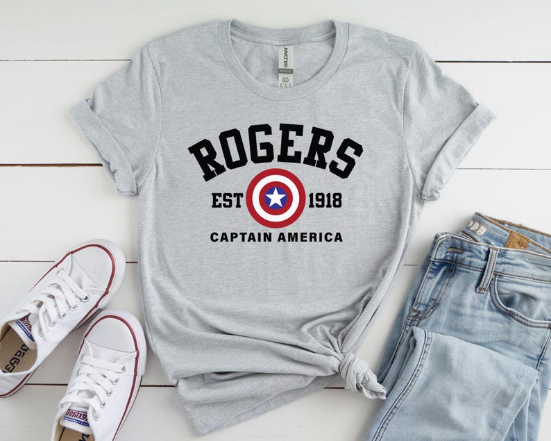 May include: A gray t-shirt with the text "ROGERS EST 1918 CAPTAIN AMERICA" in black and a Captain America shield graphic. The shirt is paired with white sneakers and blue jeans.