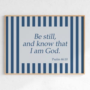Puede incluir: Un cuadro enmarcado con un marco de madera clara. La obra presenta el texto "Be still, and know that I am God." en azul sobre un fondo gris claro. El fondo tiene un patrón de rayas azules y blancas.