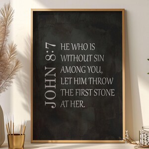 May include: A rectangular wall art print in a thin gold frame featuring a dark charcoal gray background with a watercolor texture. The text in white reads: JOHN 8:7 HE WHO IS WITHOUT SIN AMONG YOU, LET HIM THROW THE FIRST STONE AT HER.