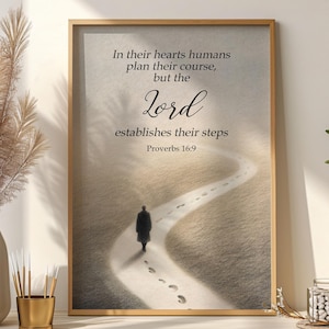 May include: Framed print with a gold-colored wooden frame. The artwork features a person walking on a path with footprints, with the text "In their hearts humans plan their course, but the Lord establishes their steps Proverbs 16:9."