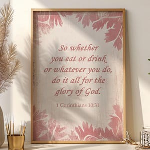 May include: A framed print with a gold-coloured frame. The print features the text "So whether you eat or drink or whatever you do, do it all for the glory of God. 1 Corinthians 10:31" in a soft, pink font. Pink floral accents frame the text.
