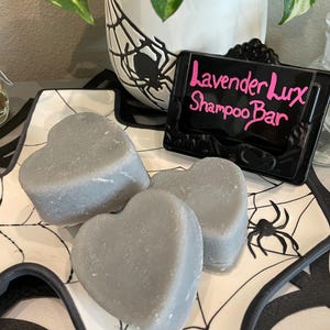 May include: Three gray heart-shaped shampoo bars on a white plate with a black spiderweb design. A black sign with the text "Lavender Lux Shampoo Bar" in pink lettering is next to the bars. A white pot with a spiderweb and spider design is in the background.