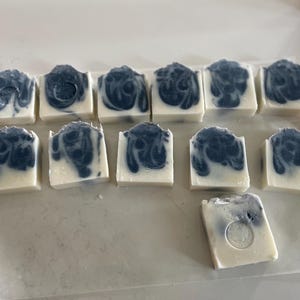 May include: A collection of handmade soaps with a white base and a dark blue, swirled design. The soaps are rectangular and arranged on a white surface. The soaps have a decorative, artistic appearance.